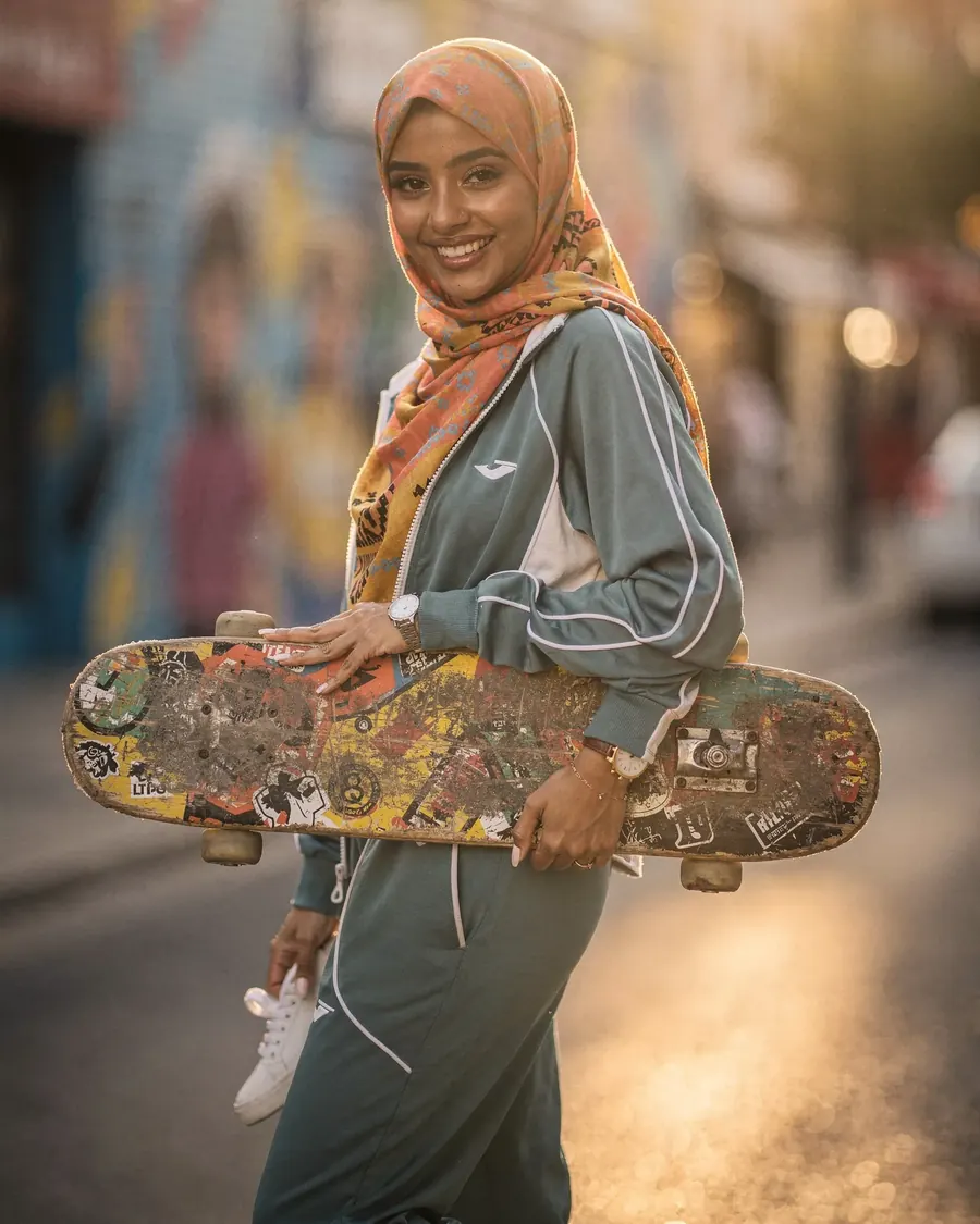 Lachende jonge vrouw met een skateboard in een zonnige straat vol muurschilderingen.
