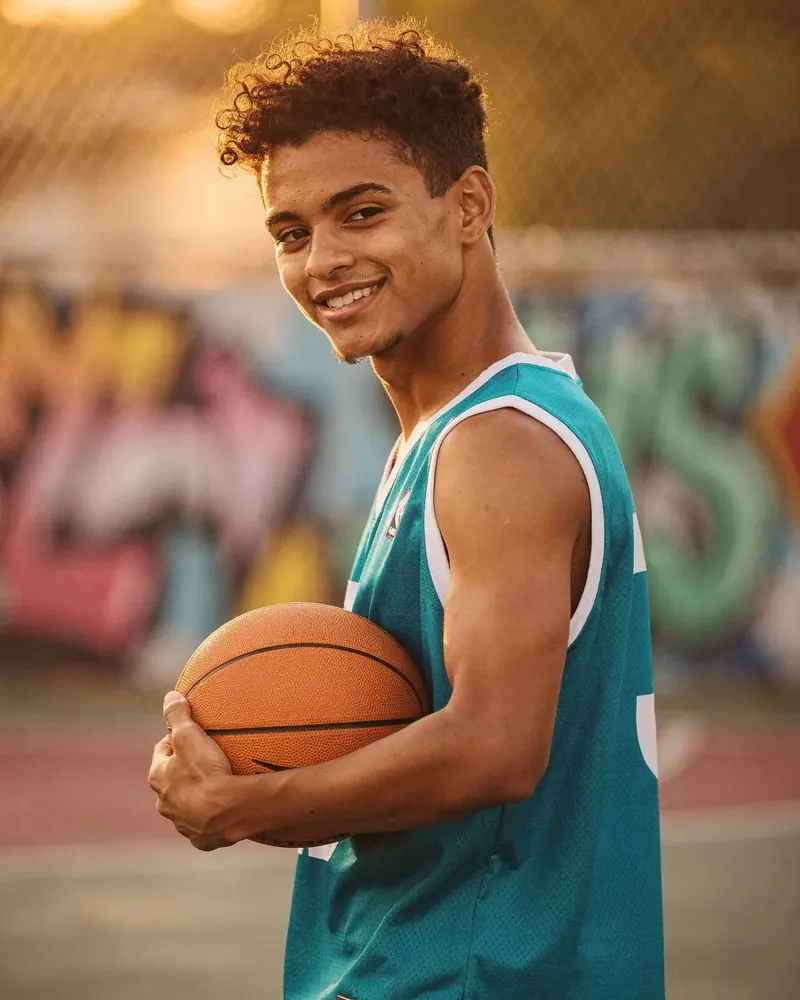 Jongeman met een basketbal lacht in de avondzon voor een graffitimuur.
