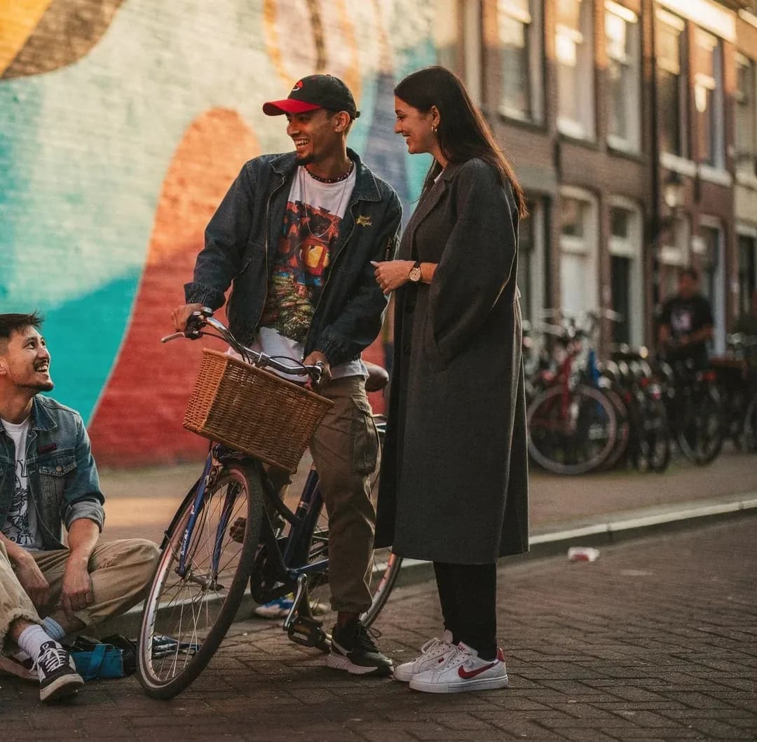 Twee vrienden met een fiets lachen voor een kleurrijke muurschildering, een vriend zit ernaast op de stoep.